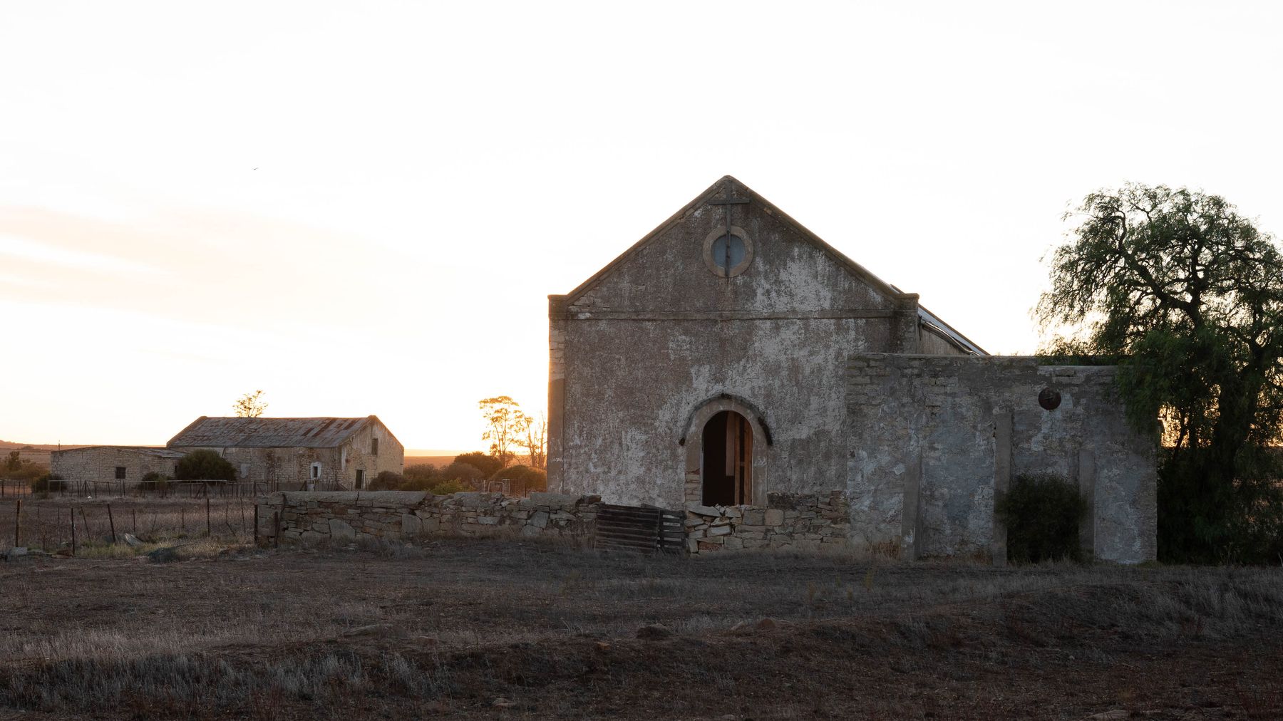 The original Nieuwoudtville church, a weathered stone building with a gable and round window, with a farmstead wall beside it in evening light