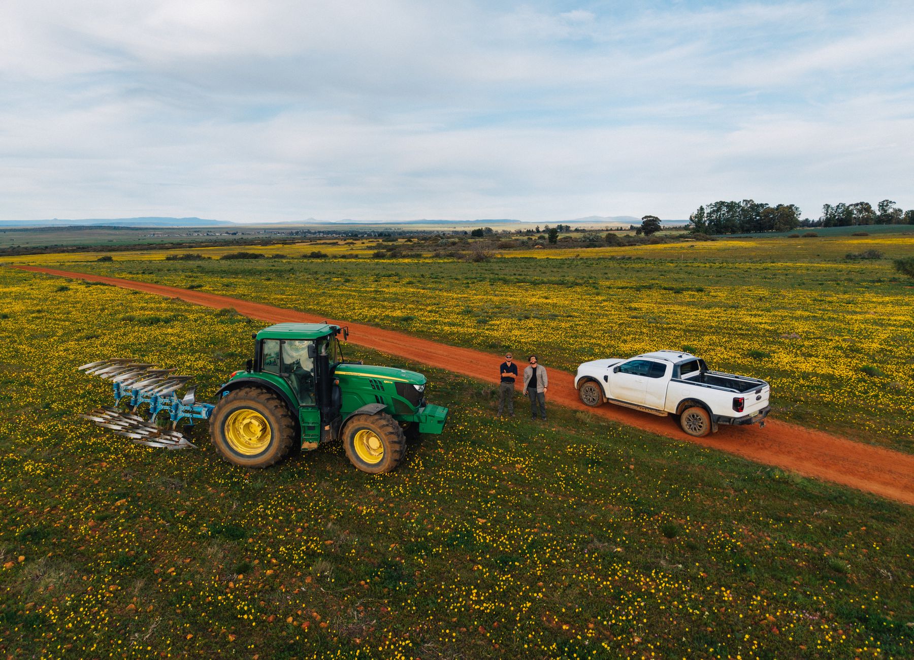 A tractor and a bakkie on a farm road through yellow flowers, from the air